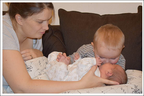 A toddler interacting with a baby that is places safely on a nursing pillow. Mom is close by to support the baby.